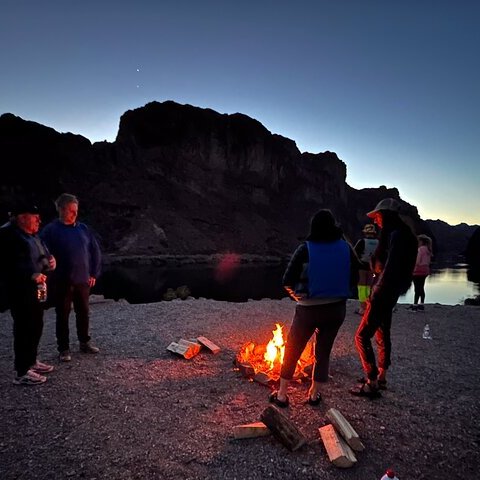 People gathered around a small campfire by a lake at dusk, with mountains silhouetted in the background.