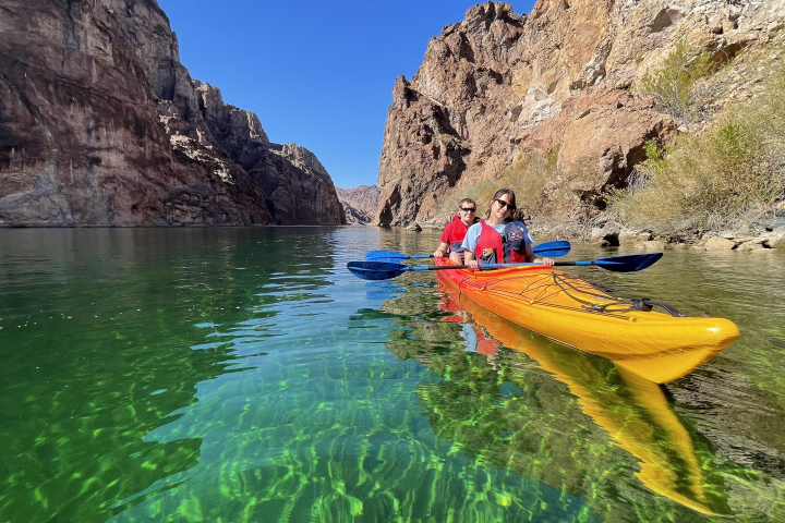 Two people kayaking on a clear green river between tall rocky cliffs.