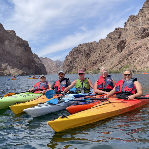 Five kayakers on a river surrounded by rocky cliffs under a blue sky.