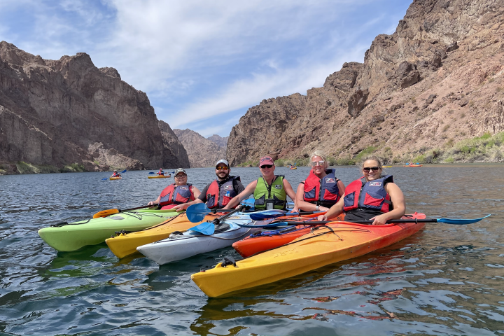Five kayakers on a river surrounded by rocky cliffs under a blue sky.