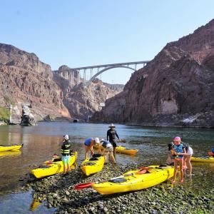 People with yellow kayaks on a rocky shore near a bridge in a canyon.