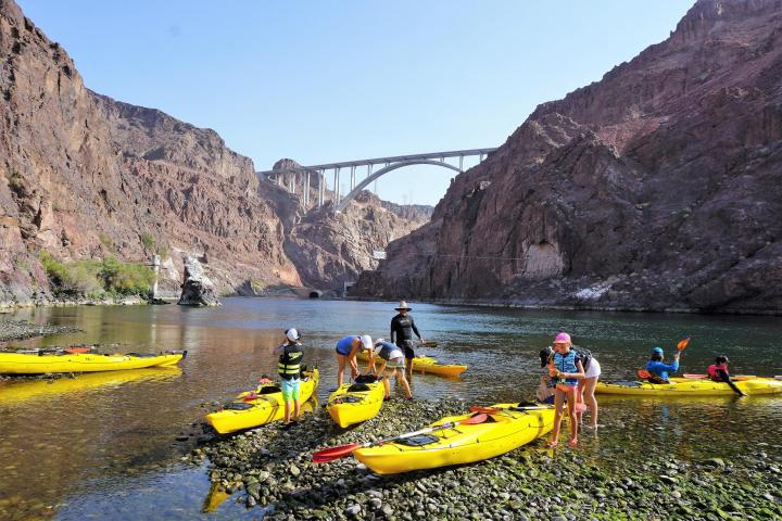 People with yellow kayaks on a rocky shore near a bridge in a canyon.