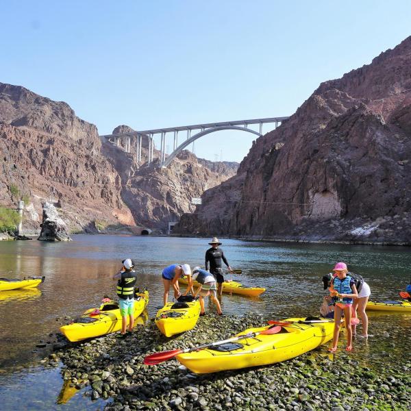 People in yellow kayaks on a rocky shore with a bridge in the background.