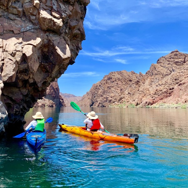 Two kayakers paddling on a clear river between rocky cliffs under a blue sky.