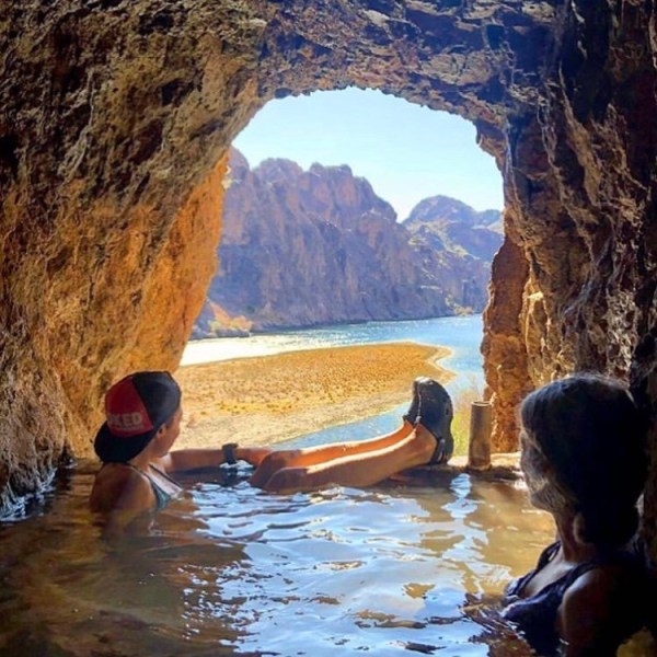 People relaxing in a cave pool overlooking a river and rocky landscape.