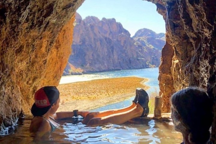 People relaxing in a cave pool overlooking a river and rocky landscape.