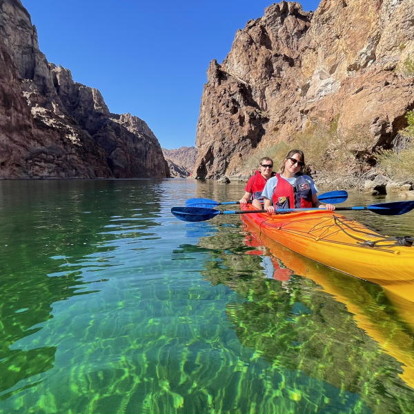 Two people kayaking on a clear river between rocky cliffs under a blue sky.
