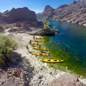 People with kayaks on a rocky riverbank surrounded by mountains.