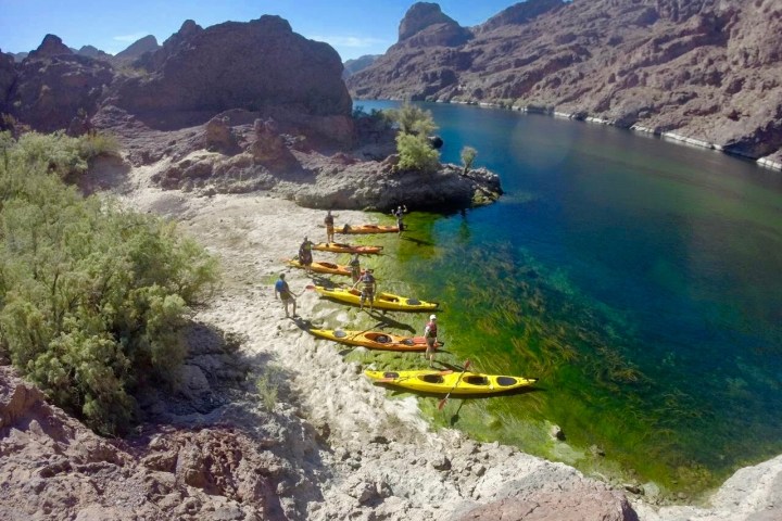 People with kayaks on a rocky riverbank surrounded by mountains.
