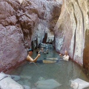 People relaxing in a narrow rocky hot spring pool, giving thumbs up.