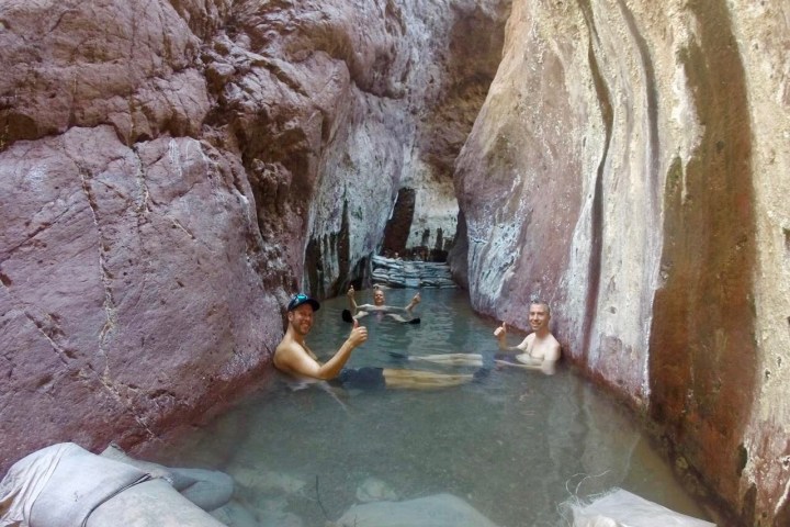 People relaxing in a narrow rocky hot spring pool, giving thumbs up.