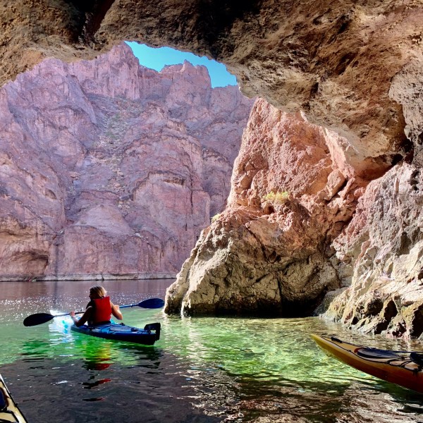 Kayakers explore a rocky cave with clear water and a mountainous backdrop.