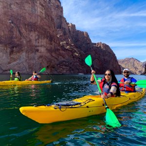 Three people kayaking on a river between rocky cliffs under a clear sky.