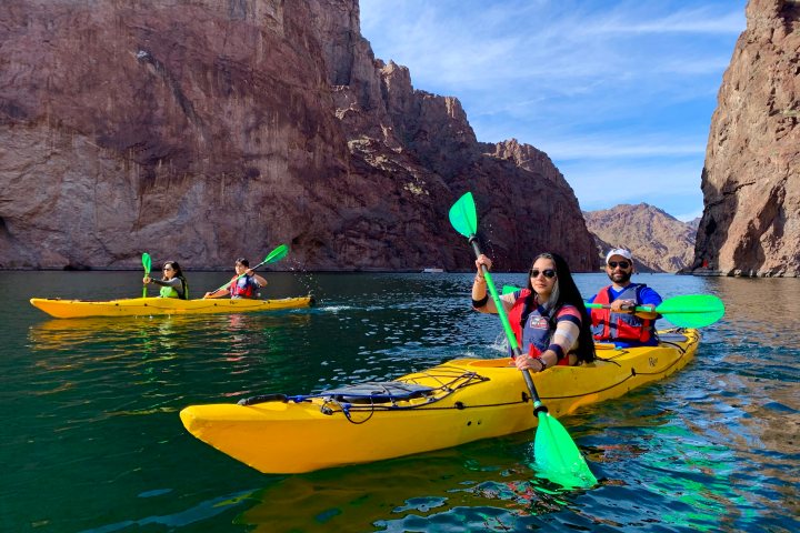 Three people kayaking on a river between rocky cliffs under a clear sky.