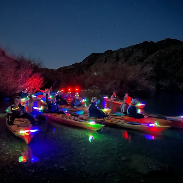 Group of kayakers with colorful lights on a nighttime river, set against silhouetted mountains.