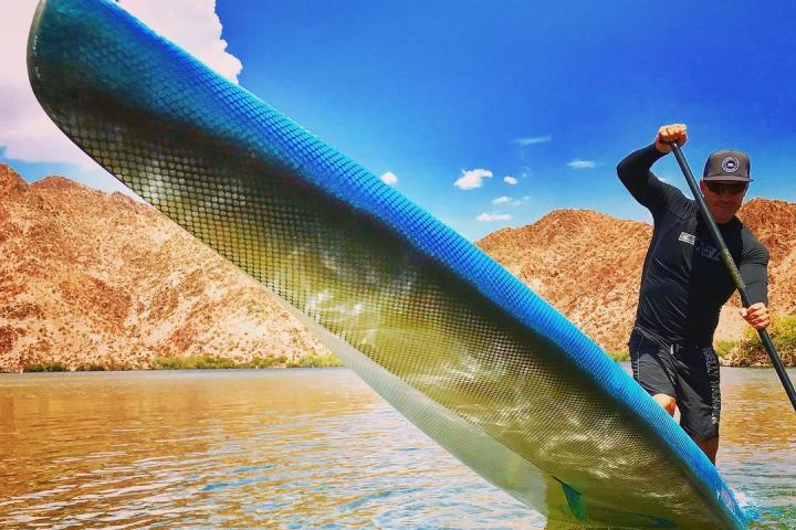 Man paddles on a lake with a paddleboard, mountains and blue sky in the background.