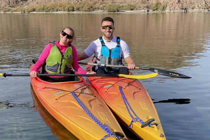 Two people kayaking on a calm lake with mountain backdrop.