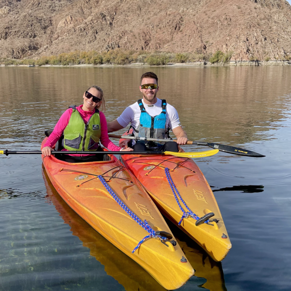 Two people kayaking on a calm lake with rocky hills in background.