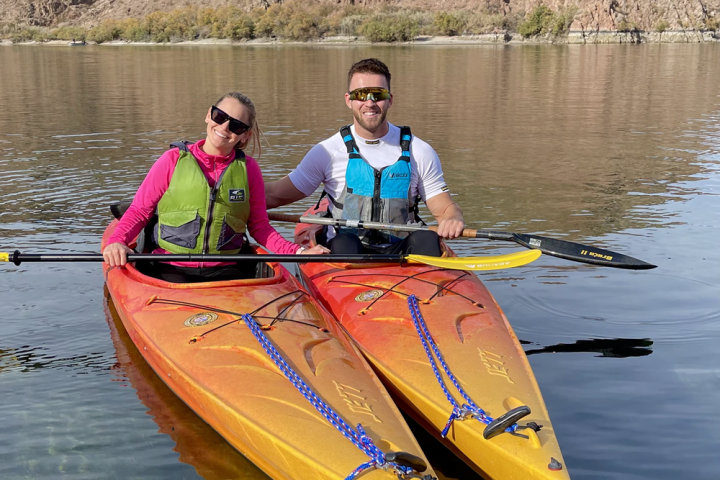 Two people kayaking on a calm lake with rocky hills in background.