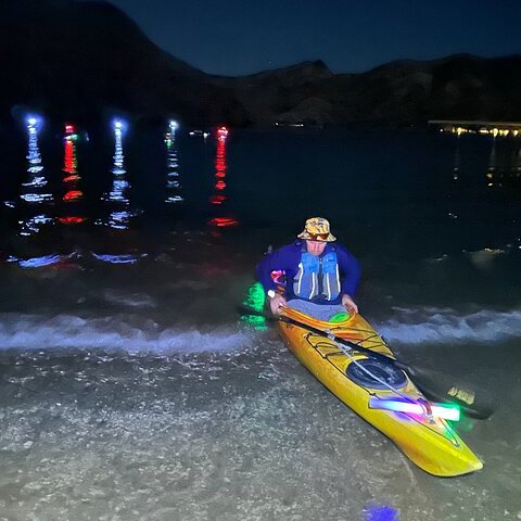 Person in a kayak with colorful lights on a dark lake, mountain silhouettes in the background.