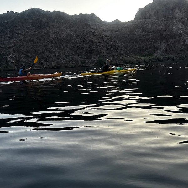 Two kayakers paddle on calm water with rocky cliffs in the background at dusk.