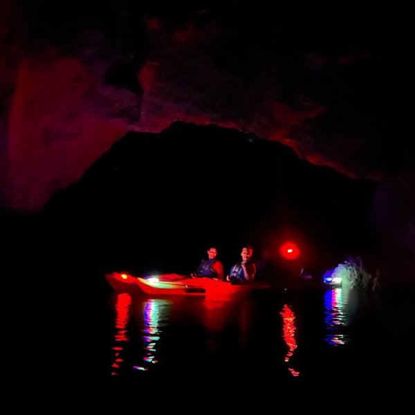 Two people kayaking in a cave with colorful lights on water.